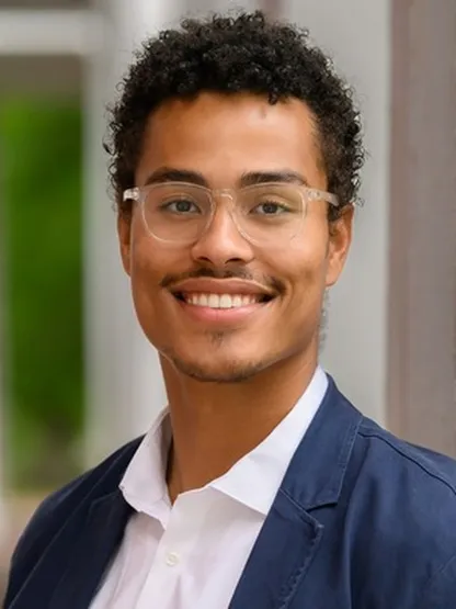 Portrait of Kai, outdoors in glasses and a jacket, looking at the camera.